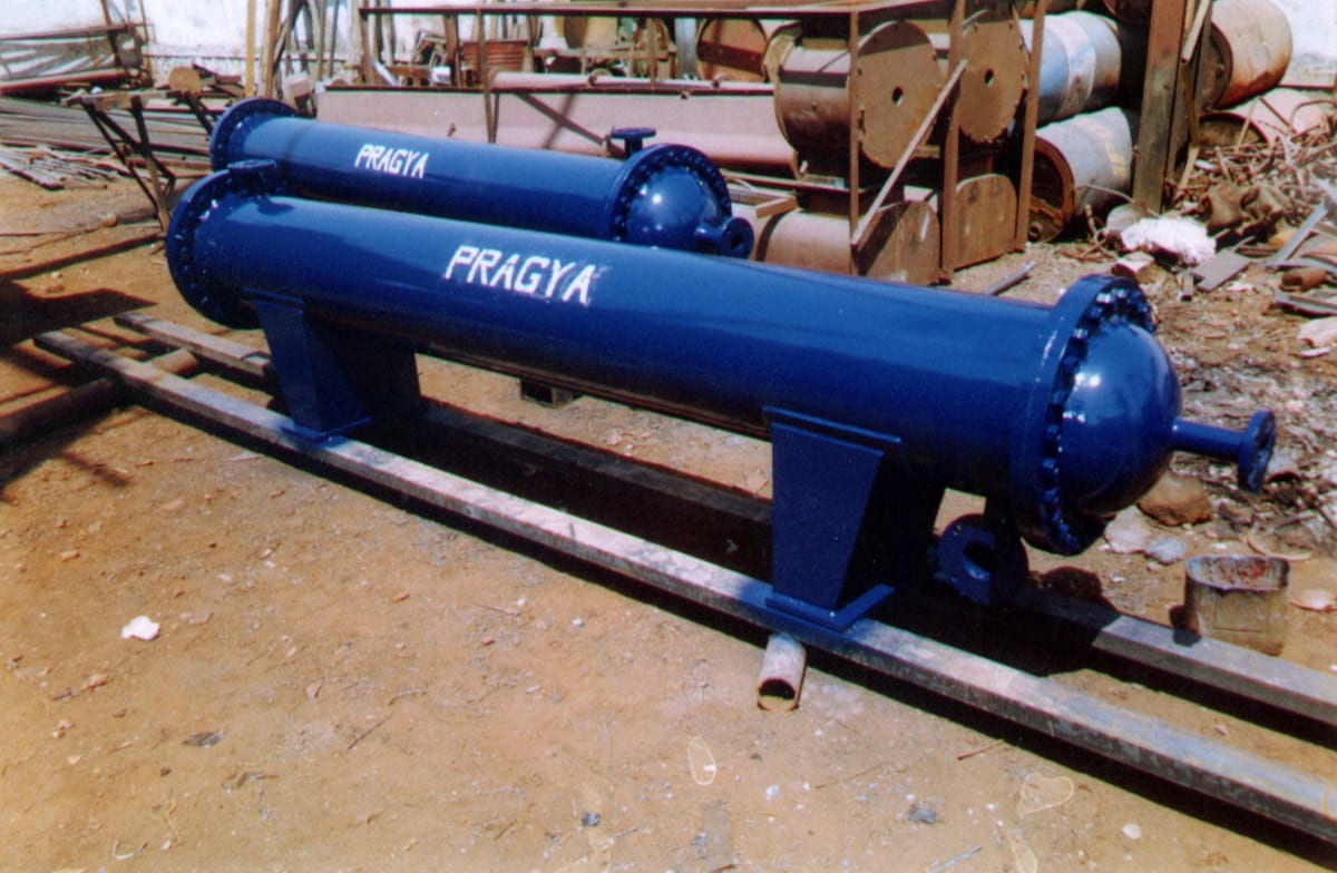 Two blue shell and tube heat exchangers lettered PRAGYA, bolted end covers and branch flanges, standing on rails in the yard