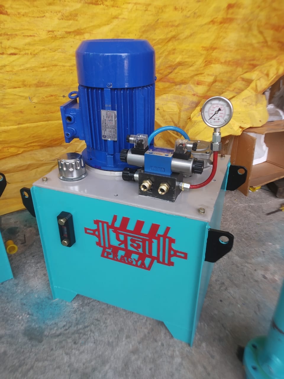 Turquoise hydraulic power pack carrying the Pragya mark, blue motor and pump on the tank top with a valve block and pressure gauge